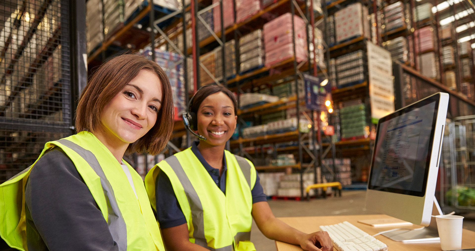 two women workers in safety vests smiling and working on a computer in the warehouse